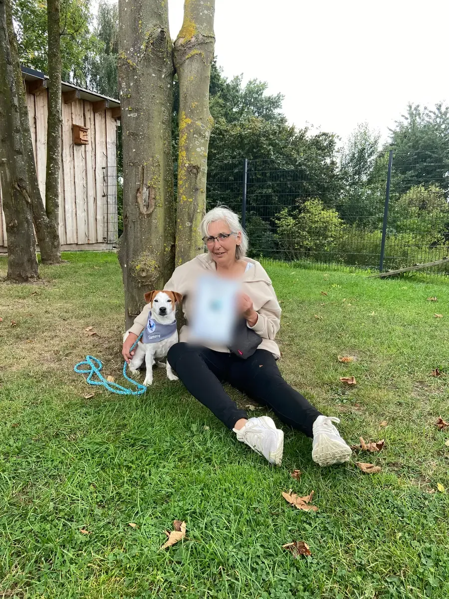 Bettina S. sitzt mit Urkunde neben ihrem Jack Russell Sammy unter einem Baum